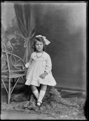 Studio portrait of an unidentified girl wearing a bow ribbon in her hair sitting on a rug covered box next to a cane chair, possibly Christchurch district