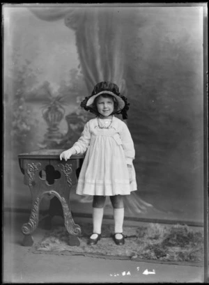 Studio portrait of an unidentified girl wearing a dress with embroidery on the front, with a ribbon tied hat and gloves, possibly Christchurch district