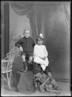 Studio portrait of an unidentified family of a boy and a girl with a bow ribbon in her hair, showing the girl sitting on a covered chair and the boy sitting on the arm rest of the chair and leaning on a cane chair next to him, possibly Christchurch district