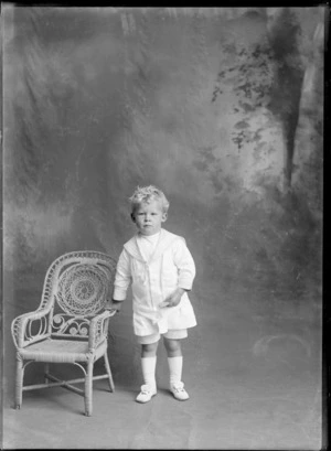 Studio portrait of an unidentified boy dressed in a white sailor suit, with long white socks, white shoes, standing next to a cane chair resting his right hand on it, possibly Christchurch district