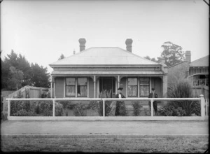 Outdoor portrait showing an unidentified elderly couple standing in the garden in front of a house on a unidentified street, showing 109 by the front door, with a enclosed fence along the street, possibly Christchurch district