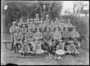 Outdoor portrait of a group of unidentified men and boys, dressed in boy scout uniforms, showing drums and drum sticks and a flag in front of some boys, in an unidentified park, possibly Christchurch district