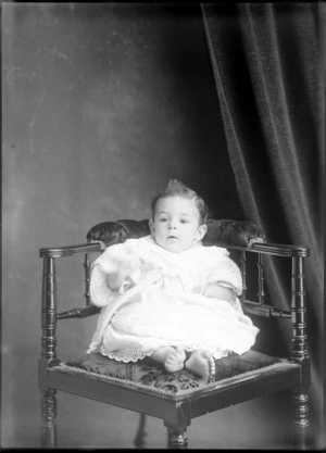 Studio portrait of an unidentified infant child dressed in a white lace gown with a matinee jacket, sitting on a wooden cushion seat, possibly Christchurch district