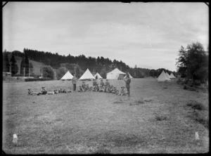 Boy Scouts camp, showing a group of unidentified scouts and leaders sitting on the ground, two of the leaders standing, one reading a book and the other leaning on his cane, with the tents in the background, in an unidentified park, possibly Christchurch district