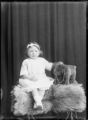 Studio portrait of unidentified girl in white dress with headband, holding circus bear on stool, probably Christchurch
