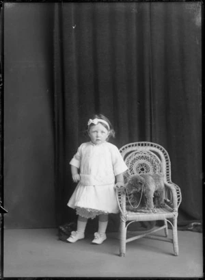 Studio portrait of unidentified young girl with toy bear on cane chair, probably Christchurch