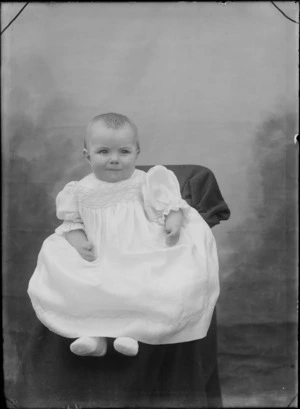 Studio portrait of an unidentified baby dressed in a embroidered gown, sitting on a highchair, possibly Christchurch district