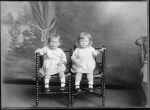 Studio portrait of unidentified [twin?] girls sitting on wooden chairs, wearing knitted dresses, possibly Christchurch district