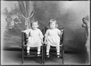 Studio portrait of unidentified [twin?] girls sitting on wooden chairs, wearing knitted dresses, possibly Christchurch district