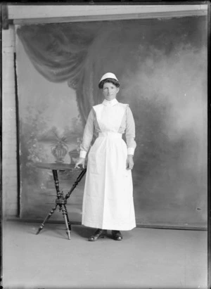 Studio portrait of an unidentified woman dressed in a nurses uniform, standing next to a wooden table resting her right hand on it, possibly Christchurch district