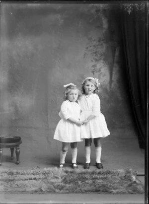 Studio portrait of two unidentified girls holding hands, wearing white dresses, long white socks and ribbons in their hair, possibly Christchurch district