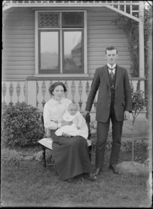 Family unidentified portrait on front lawn in front of wooden house with veranda, husband standing, wife sitting holding young baby, probably Christchurch region
