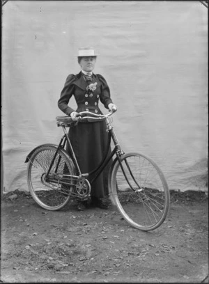 Outdoors portrait with false backdrop, an unidentified woman in dark double breasted jacket with large white buttons and flowers, tie and straw hat, standing holding women's bicycle, probably Christchurch region
