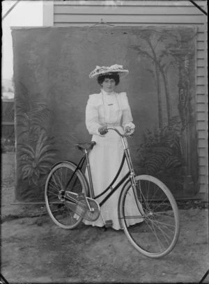 Outdoors portrait with false backdrop, unidentified woman in white lace dress and large flowery hat standing holding women's bicycle, probably Christchurch region