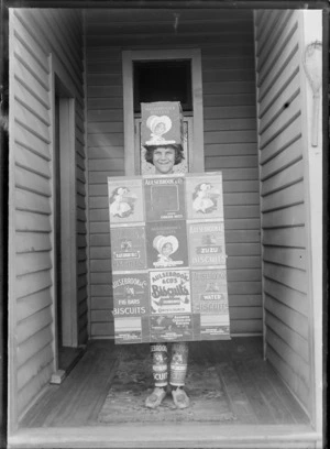 Outdoor portrait of an unidentified child in fancy dress as a 'Aulsebrook Biscuit' box, standing in the entrance of a house, possibly Christchurch district