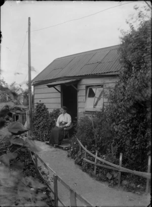 Outdoor portrait of an unidentified woman sitting on a cane chair on the stoop in front of a doorway of a house, showing the garden path in the foreground, possibly Christchurch district