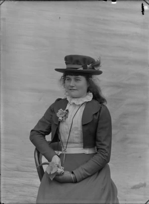 Studio portrait of an unidentified woman wearing a day dress and blouse with a lace collar, a rose attached to the front of her blouse, a hat with feathers attached to it, sitting on a wooden chair, holding a rose in her right hand, possibly Christchurch district
