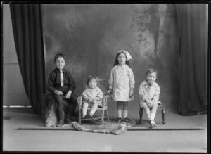 Studio family portrait of four unidentified children, older brother in white tie, black shirt, shorts and stockings and sister with hair bow, younger brother with polka dot bow, toddler sister in small cane chair, Christchurch