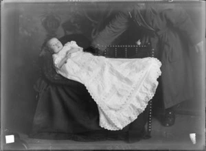 Studio portrait of unidentified baby wearing fine lace christening robe lying on small wooden couch being propped up by mother from behind, Christchurch