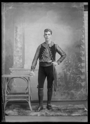Studio portrait of unidentified young man in [Eastern European?] native costume, jacket and large collar with buttons, knickerbockers, waist sash with tassels, shoes, knees and cap with rosettes, Christchurch