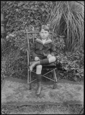 Outdoors portrait on grass in front of garden plants, unidentified young boy in sailor's outfit, sitting on bamboo chair, probably Christchurch region