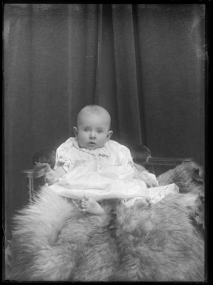 Studio portrait of unidentified baby with crocheted undergarment and cotton outer garment with bar lucky horseshoe diamond brooch, sitting on fur rug on wooden chair, Christchurch