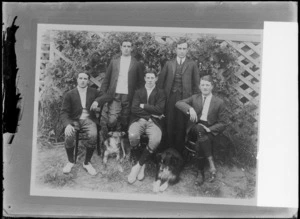 Outdoors portrait on grass in front of lattice fence, four unidentified young men in knickerbockers and high patterned socks with another man in a suit and two dogs, probably Christchurch region