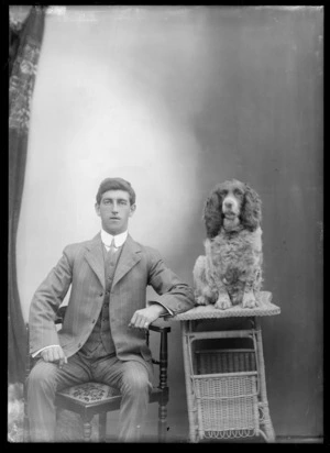 Studio portrait of unidentified young man in striped suit with double round shirt collar, sitting in chair with cocker spaniel dog on cane table, Christchurch
