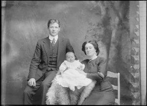 Studio family portrait, unidentified mother with high neck lace collar sitting in chair holding a baby in christening gown, and father with double round shirt collar, Christchurch