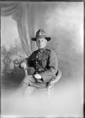 Studio portrait of the upper torso of an unidentified man dressed in army uniform holding a cane, sitting on a cane chair, possibly Christchurch district
