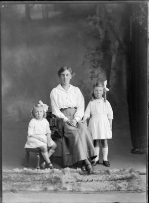 Studio portrait of members of an unidentified family, showing the woman sitting on a wooden chair, with the younger girl sitting on a stool, the older girl standing next to the woman, possibly Christchurch district