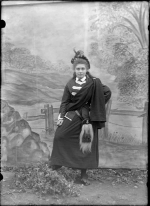 Outdoor portrait of an unidentified girl standing in front of a studio canvas backdrop, dressed in traditional Scottish costume, ankle length skirt, with a sporran hanging in the front, a cotton jacket with white panels on it, a scarf wrapped around her upper torso, draped and hanging off her left shoulder, a cap with feathers in it, possibly Christchurch district