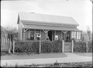 Outdoor portrait of members of an unidentified family, standing on the front porch of a house on an unidentified street, showing two boys, a man with two women, showing a hedge running along the front of the property, possibly Christchurch district