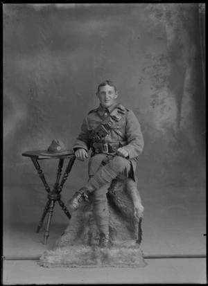 Studio portrait of unidentified New Zealand Mounted Rifles trooper with bandolier over left shoulder, ammunition waist belt, spurs and hat with silver fern badge, Christchurch
