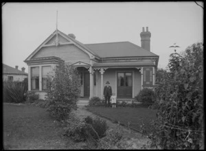 Single story wooden house with lightening rod, two chimneys and veranda, man with carnation standing with young girl on front lawn, probably Christchurch region