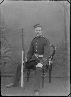 Outdoors portrait in front of false backdrop, unidentified World War One soldier with moustache and cap, 'NZE' shoulder and collar badges, sleeve insignia, crown belt buckle, with 303 rifle and bayonet, sitting in chair, probably Christchurch region