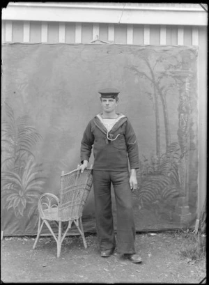 Outdoors portrait in front of false backdrop, unidentified World War One sailor with 'HMS Phoebe' on his hat and rank of 'Private', standing with cane chair, probably Christchurch region
