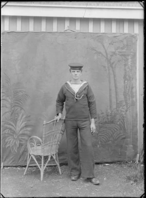 Outdoors portrait in front of false backdrop, unidentified WWI sailor with 'HMS Phoebe' on his hat and rank of 'Private', standing with cane chair, probably Christchurch region