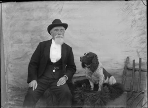 Outdoors portrait in front of false backdrop, unidentified elderly man with goatee beard and hat with wide white tie, sitting with Cocker Spaniel dog, probably Christchurch region