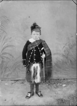 Outdoors portrait in front of false background, an unidentified young boy in Scottish clansman costume, jacket and kilt with piper's sporran, lace collar, tartan sash with brooch and Glengarry hat, Christchurch