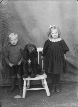 Outdoors unidentified family portrait in front of false backdrop, young girl with lace collar and boy standing with a Spaniel dog, probably Christchurch region