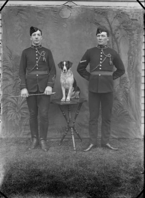 Outdoors portrait in front of false backdrop, two unidentified soldiers of the Royal Artillery in dress uniform with neck cord and tassels, forage caps, sleeve insignia, riding crop, standing with dog between them, probably Christchurch region