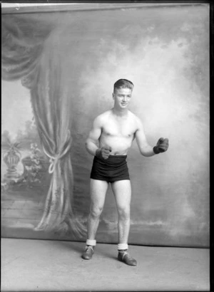 Studio portrait of unidentified young boxer in shorts, boots and woollen gloves striking a pose, Christchurch