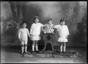 Studio portrait of four unidentified children, showing baby sitting on a carved wooden chair and the other three standing, the girls are wearing dresses and the boy shorts and jersey, all are wearing long socks below their knees and the baby is bare foot, probably Christchurch district