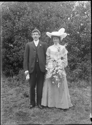 Outdoors unidentified wedding portrait in front of trees, groom with moustache and bride with large white hat holding flowers, probably Christchurch region