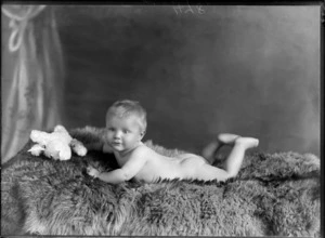 Studio portrait, unidentified baby without clothes lying front down on fur rug with teddy bear, Christchurch