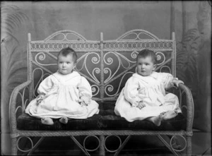 Studio portrait of two unidentified babies [twins?], wearing gowns and seated on a cane chair, probably Christchurch district