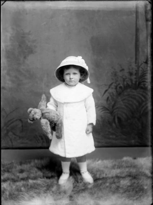 Studio portrait of unidentified small girl, wearing a coat and hat, holding a teddy bear, probably Christchurch district
