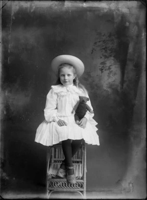 Studio portrait of unidentified young girl, seated and holding a teddy bear, probably Christchurch district