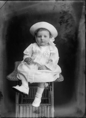 Studio portrait of unidentified small girl, wearing a hat and sitting on a cane table, probably Christchurch district